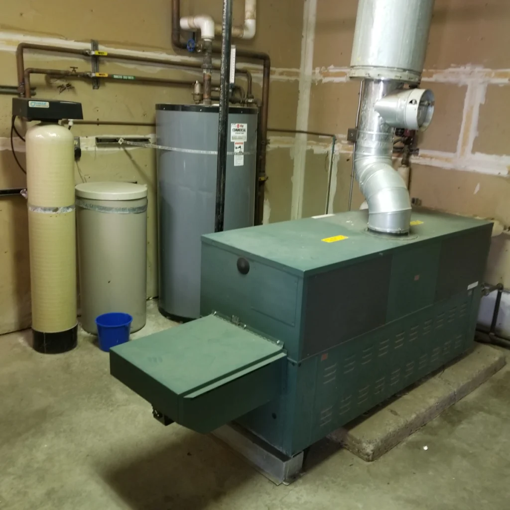 A green industrial furnace with a metal vent pipe stands in a utility room next to a water heater and a water filtration tank.