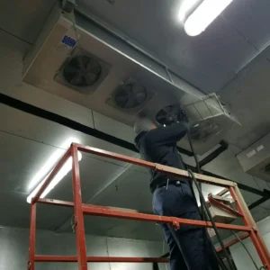 A technician on a red rolling scaffold repairs a large ceiling-mounted Bohn industrial refrigeration unit.