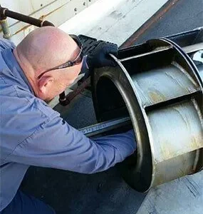 A technician in a blue uniform reaches into the circular housing of a metal industrial blower fan.