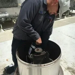 A technician in a grey work shirt and cap repairs a large industrial fan unit on a rooftop.