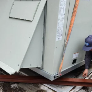 Two technicians on a roof use a metal beam to lever a large commercial HVAC unit into position.