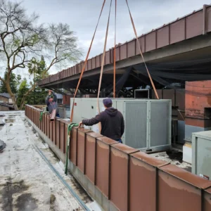 Two workers on a flat roof guide a large HVAC unit being lowered by a crane.