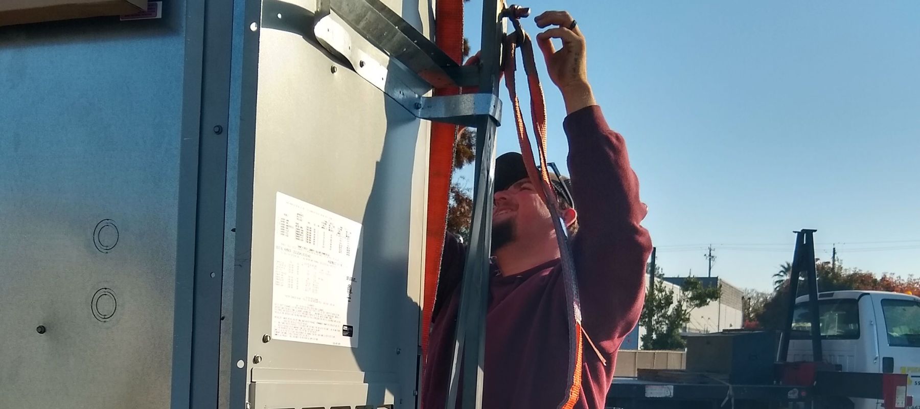 A worker secures a large metal HVAC unit onto a flatbed truck using a heavy-duty orange strap.
