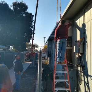 A technician on a ladder works on an electrical unit on the side of a building while a second person watches from below.