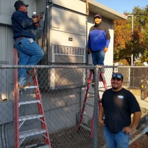 Three technicians work on installing or servicing a large Bard HVAC unit mounted to the side of a building, with two men standing on ladders and one standing on the ground.