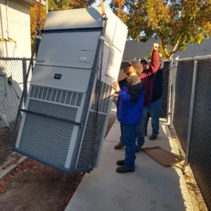 Two workers guide a large, suspended Bard HVAC unit being lowered onto a sidewalk.