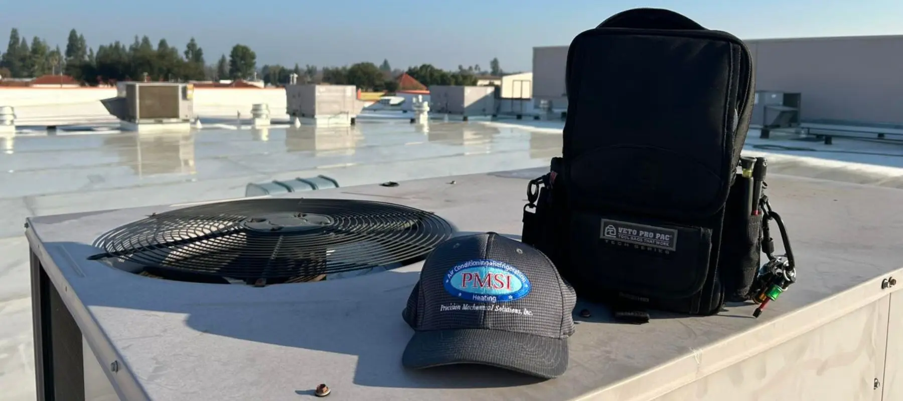 A black tool bag and a PMSI company hat rest on top of a rooftop HVAC unit under clear skies, with buildings and trees visible in the background.
