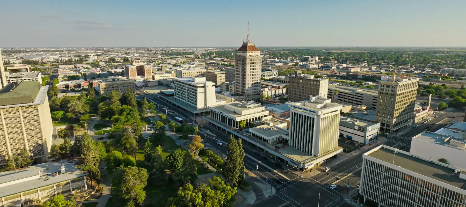Aerial view of a cityscape with a tall, pointed-roof building in the center, surrounded by other buildings, roads, and a green park with trees. The sky is clear and sunny.