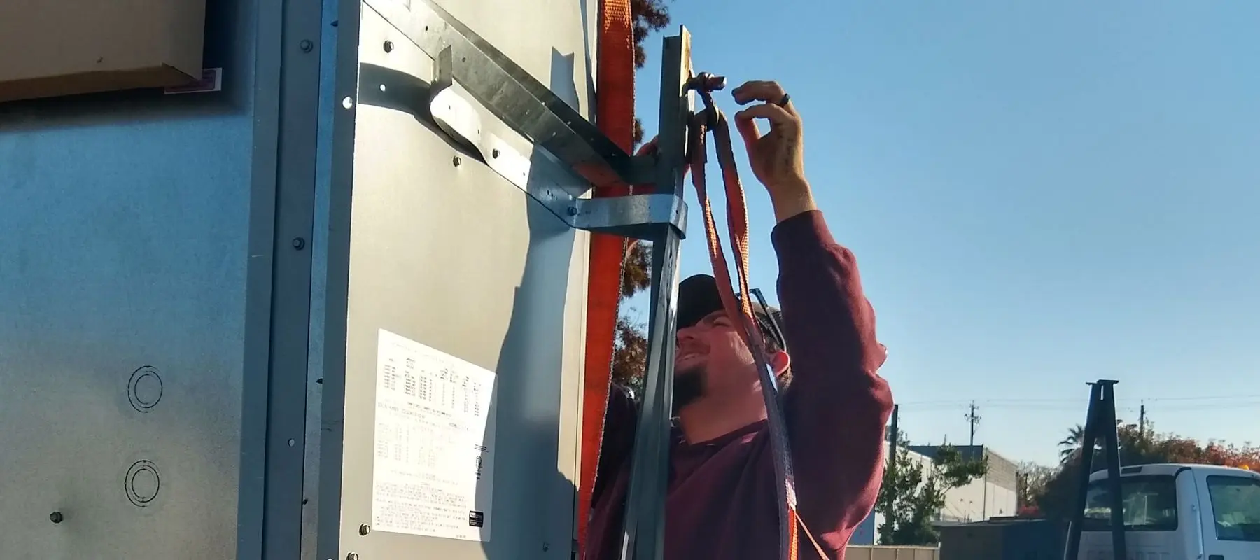 A person in a maroon shirt secures straps onto a large metal structure outdoors on a sunny day, with part of a truck and trees visible in the background.