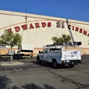 A crane lowers a large unit onto the roof of an Edwards 21 Cinemas building while a white utility truck is parked in the lot below.