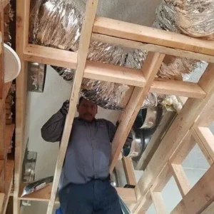 A person in a gray work shirt stands in an unfinished wooden frame while installing silver flexible air ducts.