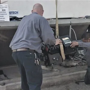 Two technicians use a hoist to install a heavy mechanical component into a large industrial cooling unit.